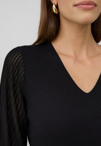 Femme portant un haut noir en V avec des manches transparentes rayées et de grandes boucles d'oreilles en or, montrant le bas du visage et le cou.