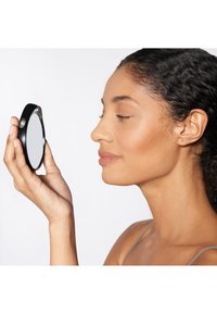 Miroir à main noir avec une surface réfléchissante ronde, tenu par une personne qui examine son visage, sur fond blanc uni.