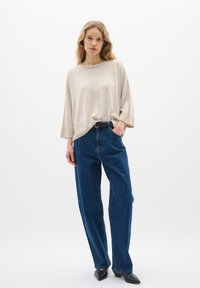 Beige oversize pulóver széles nyakkal, magas derekú kék farmernadrággal párosítva. Az öltözéket elegáns fekete hegyes cipők egészítik ki.