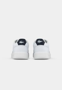Sneakers brancos com uma textura de couro suave, apresentando um tabuleiro de calcanhar em azul escuro e um detalhe de logótipo. Sola de borracha, design de bico redondo.