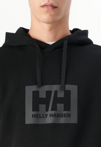 Černá mikina s kapucí, na které je velké šedé obdélníkové logo s nápisy "HH" a "HELLE HANSEN". Má kapuci se šňůrkou a strukturovanou látku.
