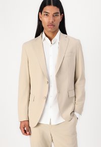 Giacca beige in tessuto leggero, caratterizzata da un design a un bottone, rever a punta e tasche frontali. Indossata sopra una camicia bianca.