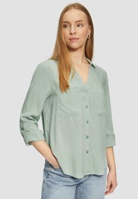 Lichtgroen, knoopshirt met V-hals, twee borstzakken, opgerolde mouwen en een gladde stoftextuur. Gecombineerd met blauwe spijkerbroeken.