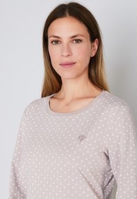 Donna con lunghi capelli castano chiaro che indossa un top a fantasia mauve chiaro con un piccolo logo "LOOP" sul petto, in piedi davanti a uno sfondo bianco.