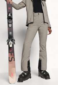 Echipamentul de schi include pantaloni impermeabili de culoare bej deschis, bocanci de schi negri și o schi cu un design în două nuanțe și legături. Textură netedă și materiale durabile.