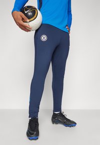 Granatowe spodenki sportowe z logo Chelsea FC, wykonane z elastycznego materiału, w zestawieniu z czarnymi korkami piłkarskimi i trzymającymi czarno-białą piłkę nożną.