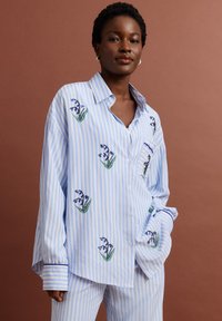 Mujer con camisa y pantalones oversize a rayas azules y blancas con bordado floral azul, de pie sobre un fondo marrón.