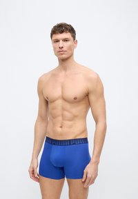 Μυώδης άνδρας που στέκεται μπροστά σε απλό φόντο, φορώντας μπλε ανδρικά εσώρουχα τύπου μπόξερ με μαύρη ζώνη με σχέδια.