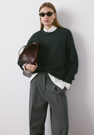 Mujer con gafas de sol negras, suéter verde oscuro sobre camisa blanca, pantalones anchos grises, sosteniendo un bolso de cuero marrón sobre fondo blanco.