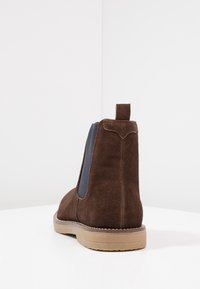 Braune Wildleder-Stiefelette mit elastischen Seitenpaneelen, einer Zuglasche auf der Rückseite und einer Gummisohle. Glatte Textur und klassische Designelemente.