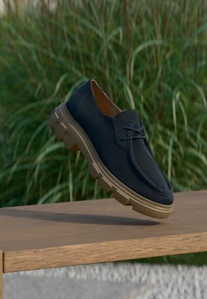 Navyblå snøresko med tyk beige sål, svævende over en træbænk, høj grønt græs sløret i baggrunden.