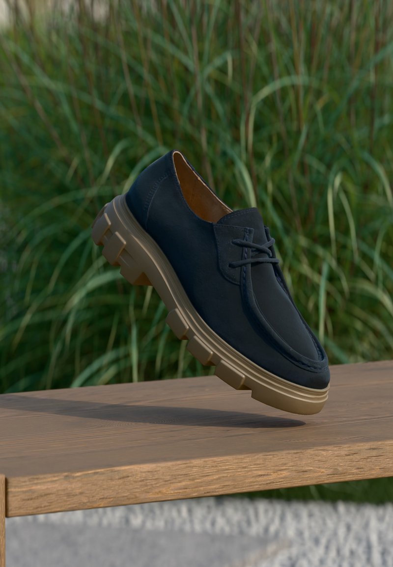 Παπουτσί από σκούρο μπλε δέρμα με κορδόνια και χοντρή μπεζ σόλα, που αιωρείται πάνω από ξύλινο παγκάκι, με ψηλό πράσινο γρασίδι φλουταρισμένο στο φόντο.