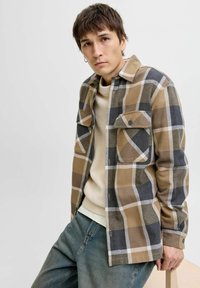 Camicia a scacchi marrone e grigia con due tasche sul petto, chiusura con bottoni e una texture morbida, indossata sopra un maglione color crema e jeans blu.