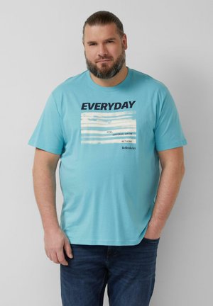Mann mit Bart, der ein hellblaues T-Shirt mit dem Schriftzug „EVERYDAY“ und Textstreifen trägt, steht mit einer Hand in der Tasche vor einem einfachen Hintergrund.