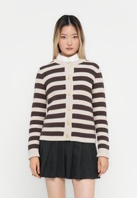 Ung kvinne iført en beige og mørk brun stripete genser med gullknapper og et plissert svart skjørt, står mot en hvit bakgrunn.