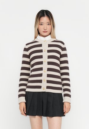 Ung kvinne iført en beige og mørk brun stripete genser med gullknapper og et plissert svart skjørt, står mot en hvit bakgrunn.