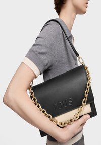 Persona con camisa de manga corta gris sosteniendo un bolso de hombro negro de cuero TOUS con cadena dorada, contra un fondo blanco.