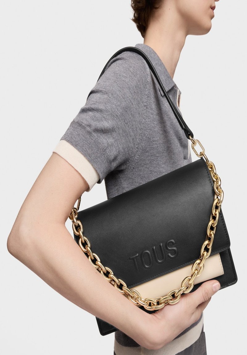 Persona con camisa de manga corta gris sosteniendo un bolso de hombro negro de cuero TOUS con cadena dorada, contra un fondo blanco.