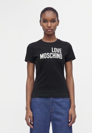 Giovane donna dalla pelle scura e capelli raccolti indossa una t-shirt nera Love Moschino e jeans blu, in piedi su sfondo bianco.