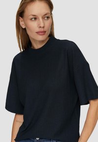 Granatowy top z krótkim rękawem i wysokim dekoltem, o miękkiej fakturze, luźnym kroju oraz widocznymi detalami przeszycia, zestawiony z dżinsami.