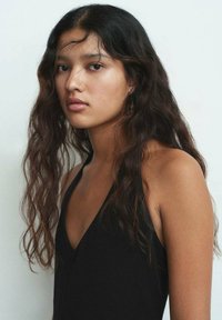 Prenda negra sin mangas con un profundo escote en forma de V, mostrando cabello oscuro ondulado hasta los hombros y un cutis suave contra un fondo claro.