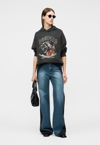 Γυναίκα με μαύρη φούτερ με στάμπα πάνθηρα, μπλε τζιν παντελόνι φαρδιάς γραμμής, μαύρα γυαλιά ηλίου, κρατώντας μαύρη τσάντα.