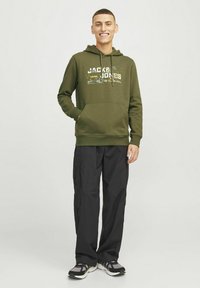 Groene hoodie met voorzak en trekkoorden, voorzien van witte en gele tekst, in combinatie met zwarte cargobroeken en meerkleurige sneakers.