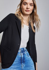 Blazer nero con texture, revers a punta, chiusura con un bottone e tasche anteriori, indossato sopra una camicia bianca e pantaloni denim blu.