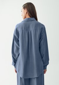 Camicia blu a maniche lunghe con colletto, texture liscia, lucentezza sottile e orlo curvo. Presenta una piega sulla schiena per una maggiore libertà di movimento.