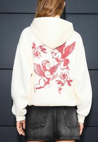 Cream hoodie with a large red graphic of cherubs and roses on the back. Features a drawstring hood and loose fit. Paired with a black denim skirt.