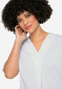 Witte, loszittende blouse met een V-hals en korte mouwen. De stof lijkt glad met een subtiele textuur en stiksels bij de halslijn.