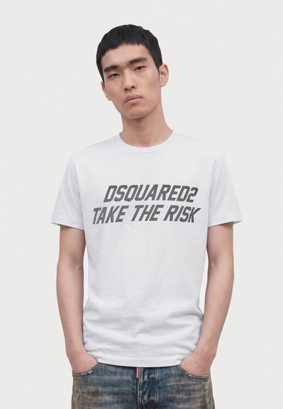 Jeune homme aux cheveux noirs courts porte un t-shirt blanc avec le texte "DSQUARED2 TAKE THE RISK", mains dans les poches, sur un fond clair uni.