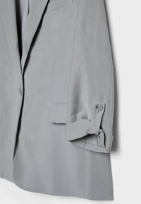 Blazer gris clair à un bouton, poche à rabat et manches retroussées maintenues par une patte à bouton sur un fond blanc.