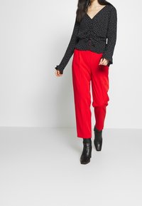 Blusa nera a manica lunga con pois bianchi, aderente in vita, abbinata a pantaloni rossi luminosi a vita alta e stivali neri alla caviglia.