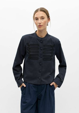 Femme à la peau claire et aux cheveux attachés porte une veste sombre avec des boucles décoratives et des boutons sur le devant, mains dans les poches d'un pantalon bleu marine.