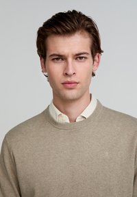Beige trui van wol, ronde hals, zachte textuur, met een subtiele logo op de borst, gelaagd over een lichte overhemd met kraag.
