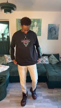 Tânăr stând în living, purtând o cămașă neagră cu mâneci lungi și un design colorat, pantaloni deschiși la culoare și cizme închise, uitându-se în jos și zâmbind.