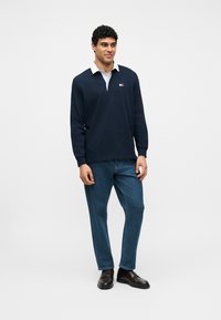 Άνδρας που φοράει μπλε μπλουζάκι ράγκμπι με λευκό γιακά, μπλε τζιν και σκούρα καφέ μοκασίνια, στέκεται μπροστά σε ένα απλό φωτεινό φόντο.