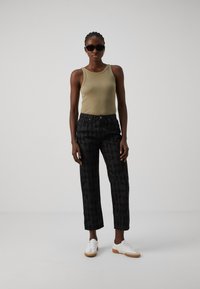 Olivgrünes geripptes Tanktop, schwarze gemusterte weitgeschnittene Jeans, weiße Sneakers mit Gummi-Sohlen, schwarze Sonnenbrille, minimaler Schmuck.