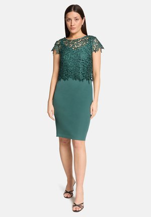 Kvinde iført en ærmeløs mørkegrøn knælængde kjole med blomsterblondeoverlay på den øverste del af kroppen og sorte stroppesandaler.