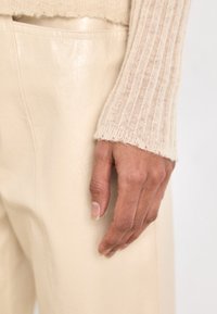 Manica di un maglione a coste beige accanto a un pantalone in pelle beige chiaro, liscio, con una tasca laterale visibile. Texture morbida e linee pulite.