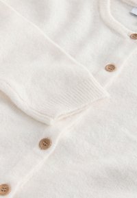 Cardigan tricoté d'un blanc cassé doux avec des boutons marron clair et des poignets côtelés, présenté en gros plan.
