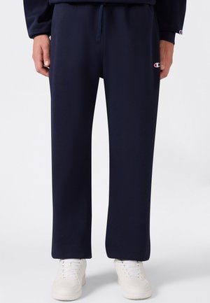 Persona che indossa pantaloni della tuta blu navy con coulisse e logo Champion sulla coscia sinistra, abbinati a scarpe da ginnastica bianche.
