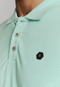Mintgroene polo met een textuurstof, voorzien van twee knopen en een zwart hexagonaal logo met gouden accenten op de borst.