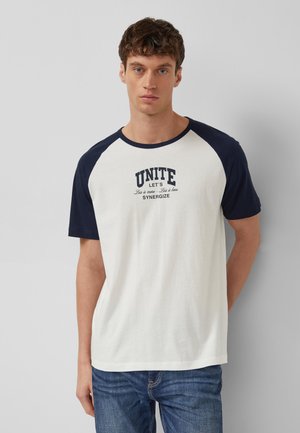 Junger Mann trägt ein weißes T-Shirt mit marineblauen Raglanärmeln und dem Aufdruck "UNITE LET'S SYNERGIZE", kombiniert mit blauen Jeans, stehend vor einem grauen Hintergrund.