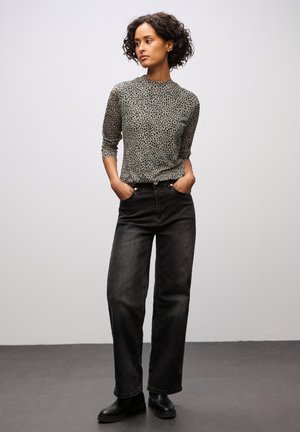 Una donna con capelli ricci che indossa una maglietta a maniche lunghe grigia con macchie, pantaloni neri a vita alta e larghi, e stivali neri alla caviglia, in piedi con le mani in tasca.