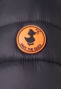 Černá péřová bunda s kulatou oranžovou gumovou nášivkou s siluetou kachny a textem "SAVE THE DUCK." Texturovaná tkanina.