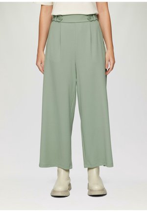 Frau trägt hochtaillierte, breite, sandgrüne Hosen mit Knopfdetails am Gürtel und off-white klobige Stiefel, steht vor einem schlichten Hintergrund.