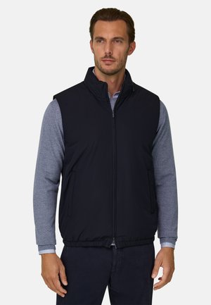 Homme portant une veste sans manches noire à fermeture éclair sur une chemise grise à manches longues, debout les mains détendues le long du corps.