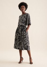 Abito nero con motivo paisley bianco, maniche corte, vita elastica e tasche. Tessuto morbido e design fino al ginocchio. Sandali neri.